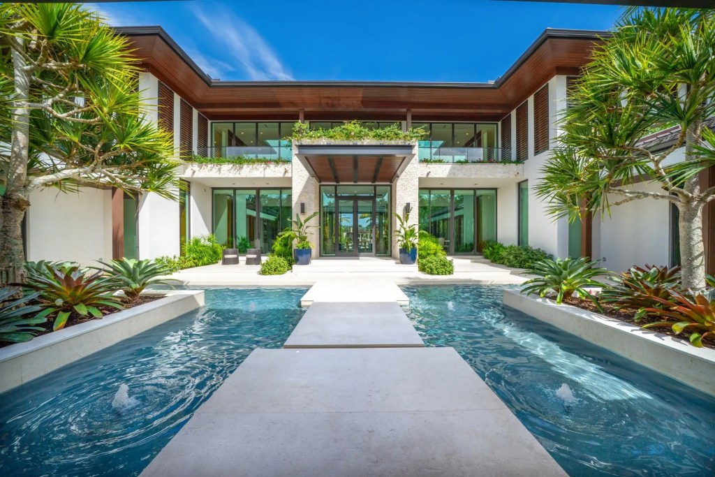 Grand contemporary waterfront estate featuring a dramatic entrance courtyard with reflecting pools, stepping-stone walkway, tropical landscaping, and expansive glass walls. Coral Gables, Florida.