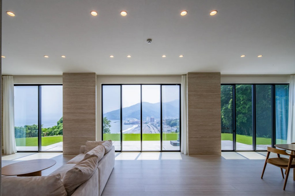 Modern open-plan living room with floor-to-ceiling glass doors framing panoramic coastal and mountain views, leading to a spacious terrace and manicured lawn. Kanagawa, Japan.