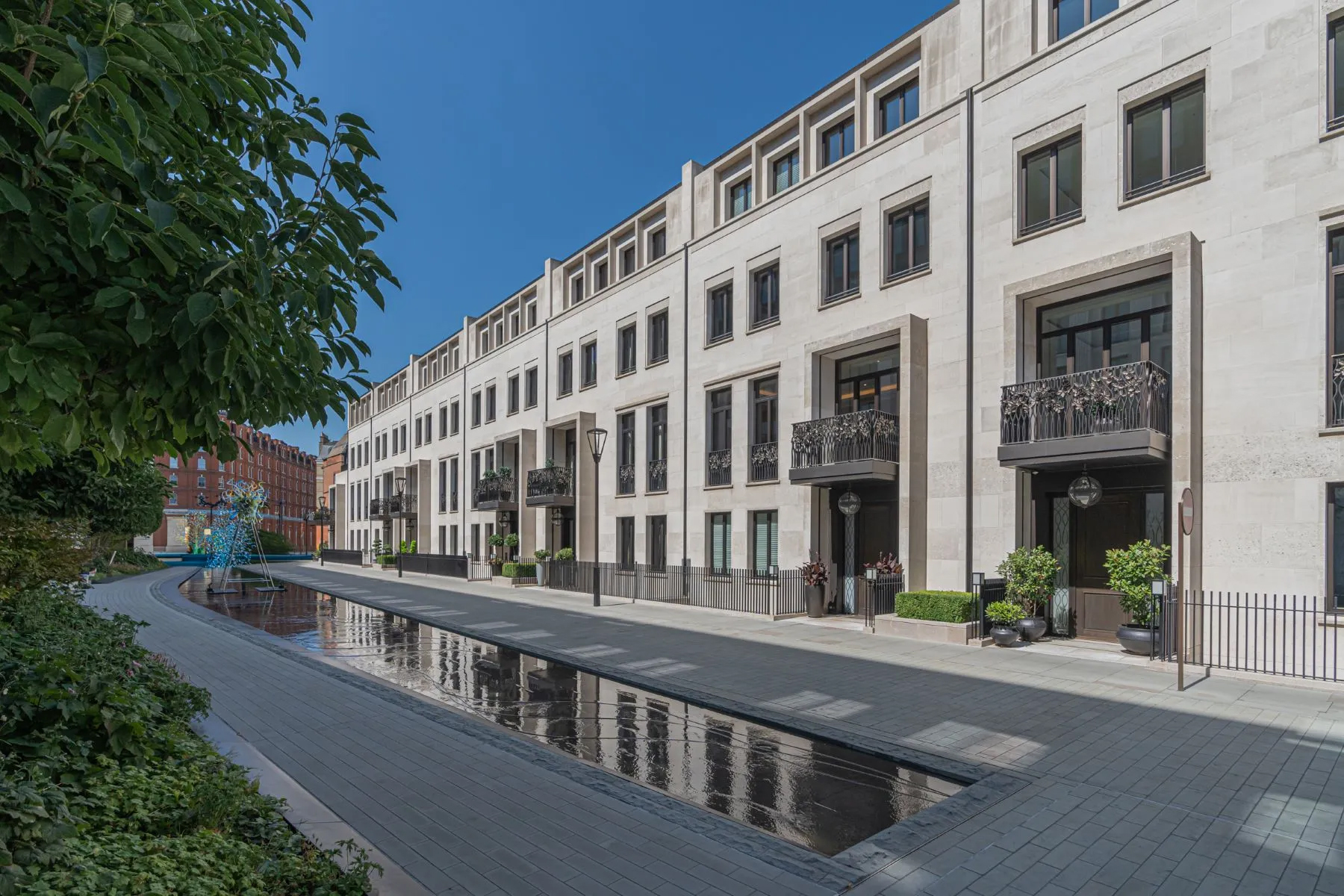 Contemporary luxury townhomes with stone façades, private balconies, and a reflective water feature along a landscaped pedestrian street. London, England.