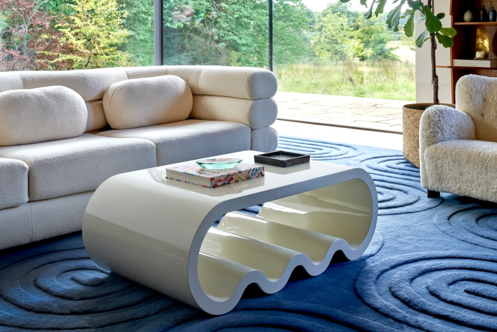 Modern living room featuring a white curved sofa, a sculptural glossy white coffee table, and a textured blue rug, with floor-to-ceiling windows overlooking a green landscape.