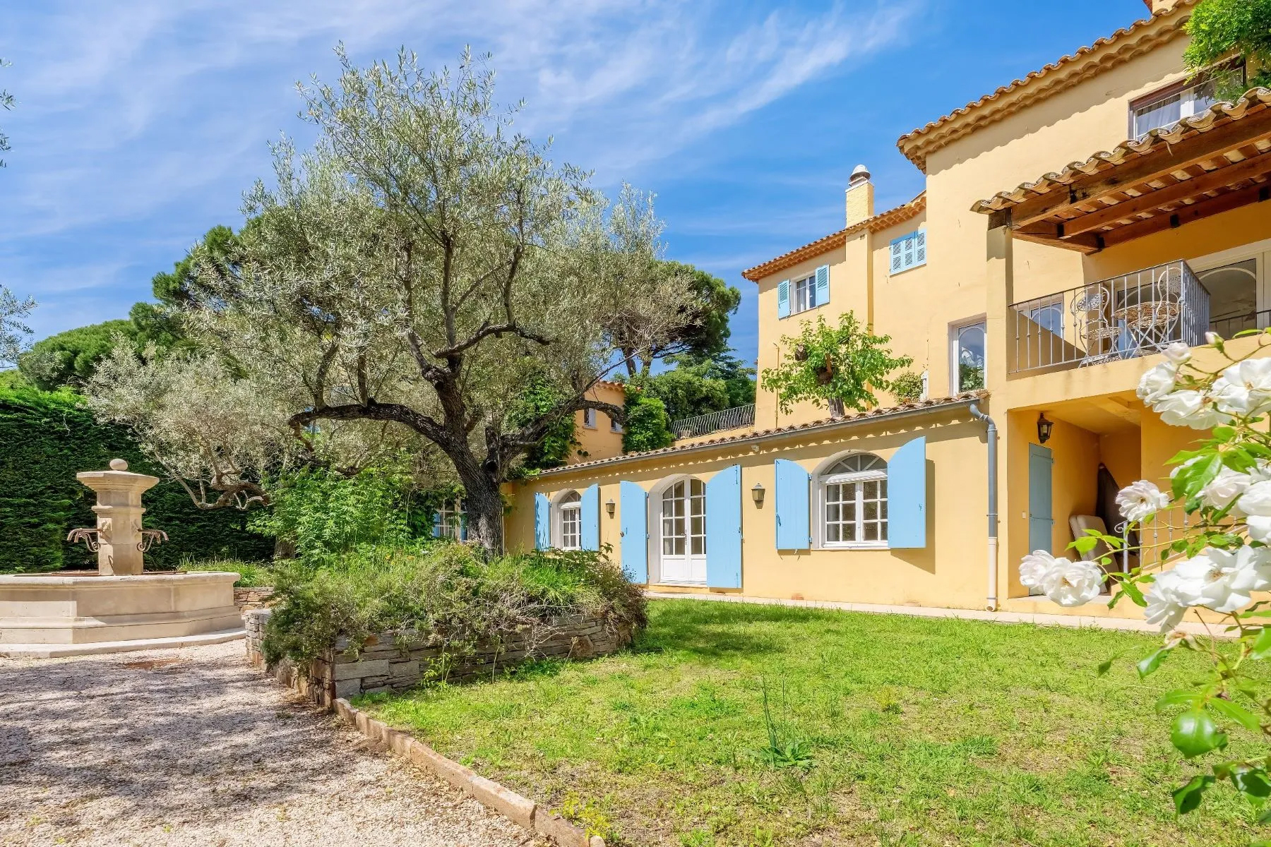 Mediterranean-style villa with pale yellow walls and blue shutters, surrounded by mature olive trees, flowering plants, and a stone fountain in a sunlit garden. Sainte Maxime, France.