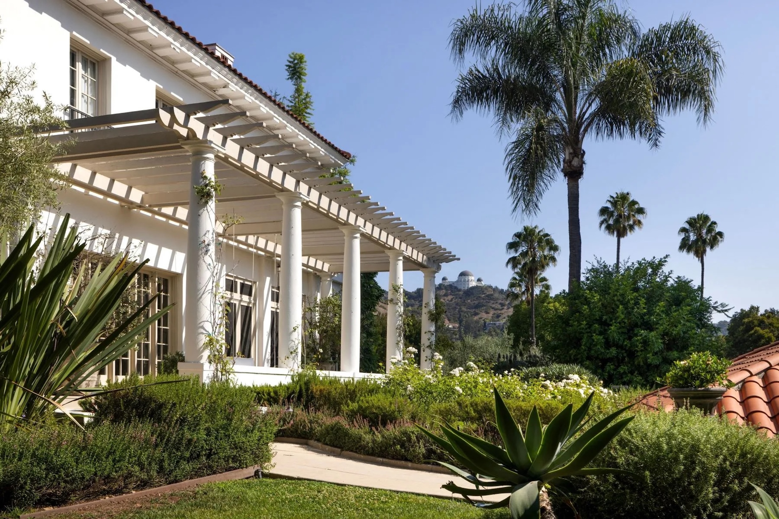 Veranda on DeMille estate surrounded by lush greenery and palm trees, Griffith Observatory in the distance