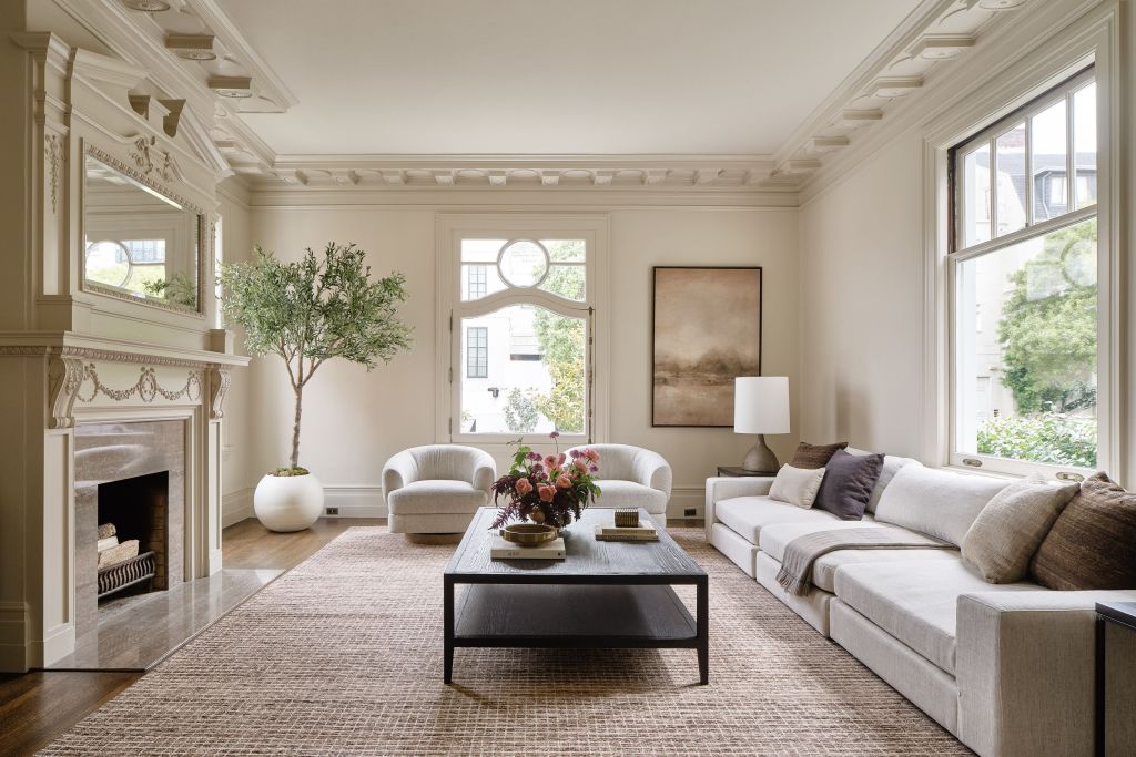 elegant white sitting room with marble fireplace with ornate wood molding, a white sofa, two chairs, a long coffee table, art on walls, plant in corner, and windows looking out to the tree-lined streets