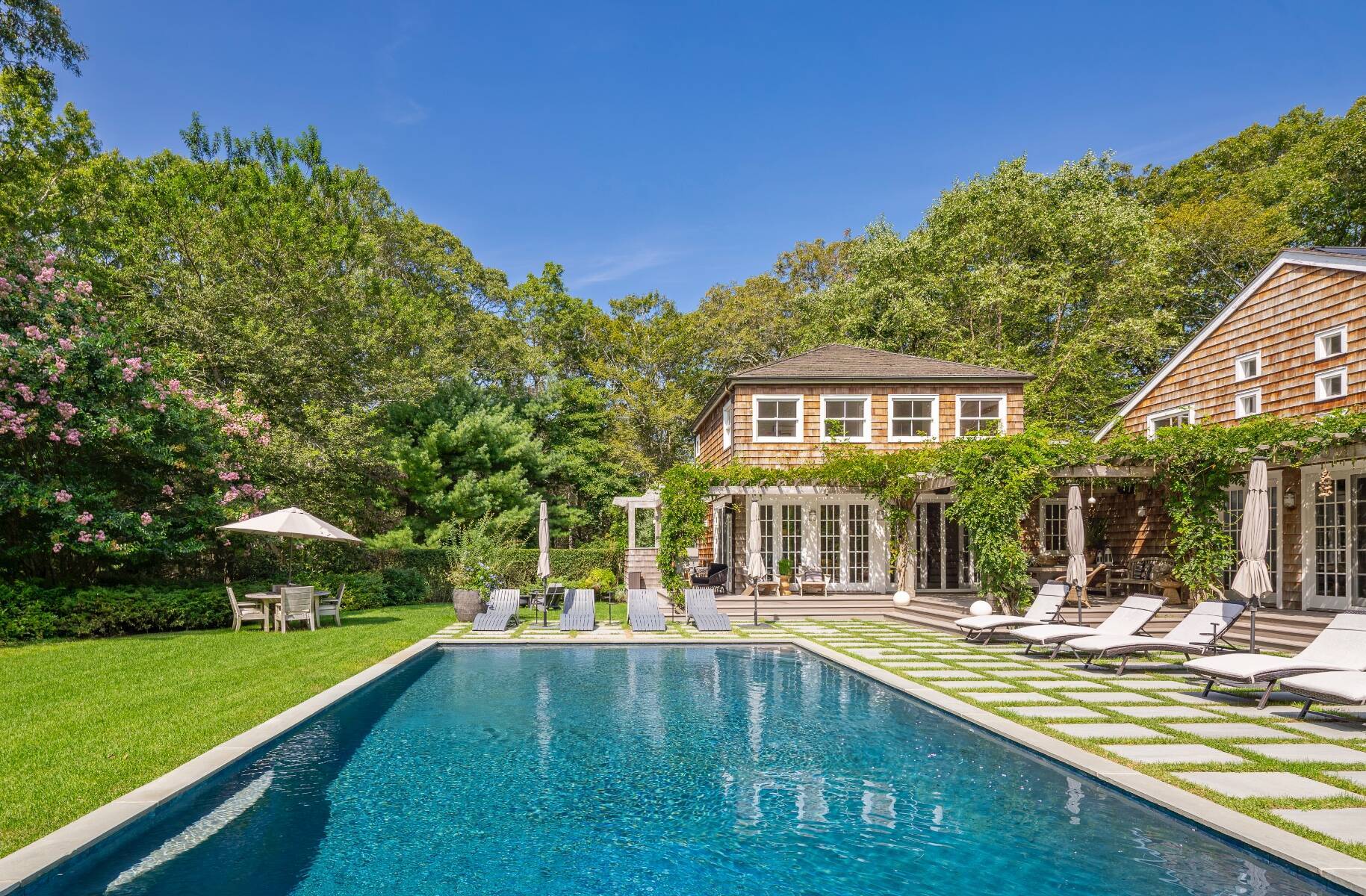 View from the pool of Amagansett, New York home exterior with shingle-style architecture and white-bordered windows and outdoor seating enveloped in lush landscaping