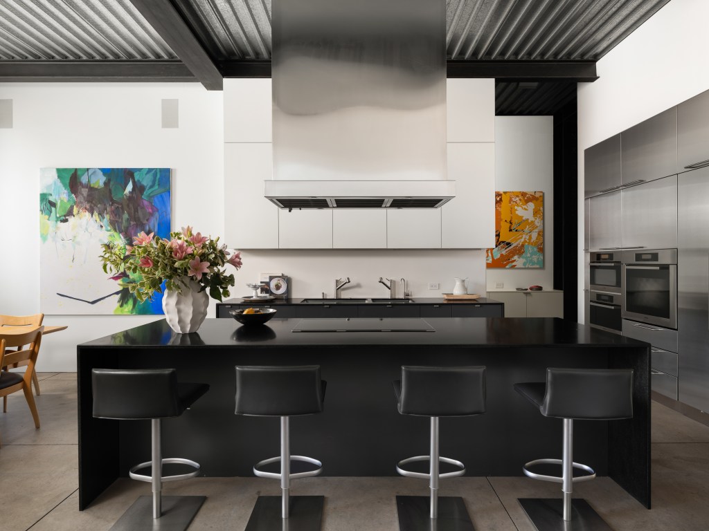 contemporary kitchen in San Francisco's Gallery House, featuring corrugated metal ceilings, black waterfall island, four leather barstools, and stainless steel vent hood. White walls in the background showcase art