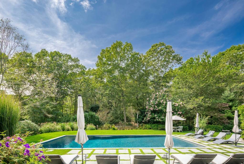 backyard of Amagansett home with towering trees, lush landscaping, and pool deck leading out to gunite pool
