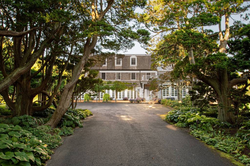 Long driveway lined with mature trees and lush landscaping leading to a large shingle-style residence with white-trimmed windows and a covered front entrance.