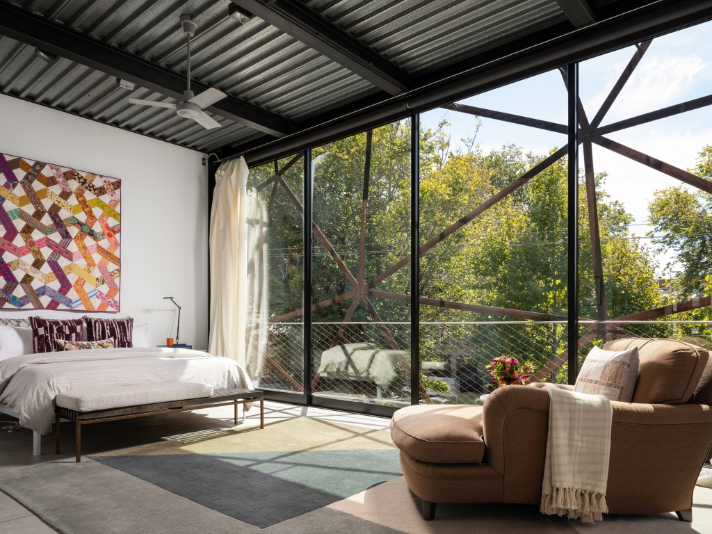 bedroom with quilted art on the wall behind the bed, chaise lounge, and artistic steel webbing on the window, overlooking treetops