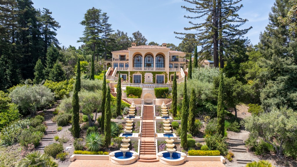 Mediterranean-style estate surrounded by mature trees and terraced gardens, featuring a grand central staircase, tiered fountains, arched windows, and formal landscaping viewed from above.