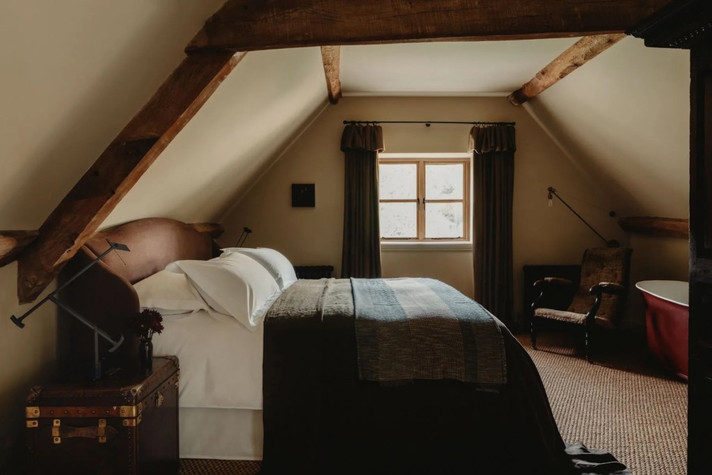 bedroom in reimagined barn in the Cotswolds, featuring exposed wood beams, cozy blankets and bedding, and soft lighting streaming from the window in the background