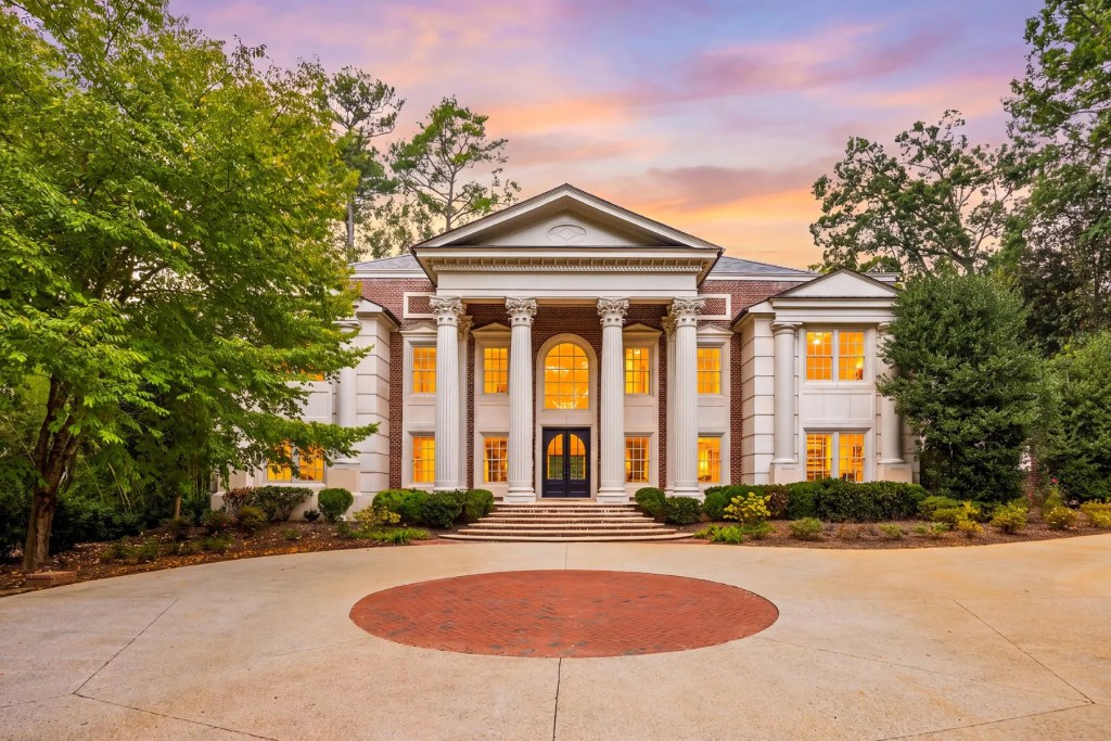 Large neoclassical estate with tall white columns and a circular driveway, surrounded by mature trees and landscaped grounds at sunset.