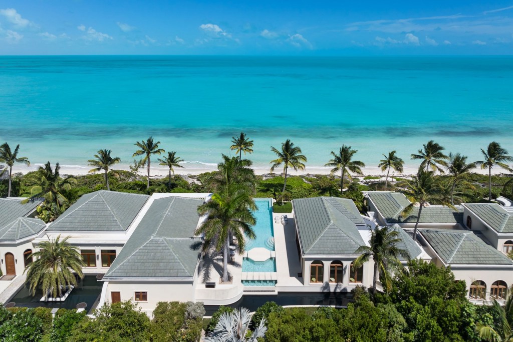 Oceanfront luxury estate with multiple pavilions, palm-lined grounds, and a central swimming pool overlooking a white-sand beach and turquoise water.