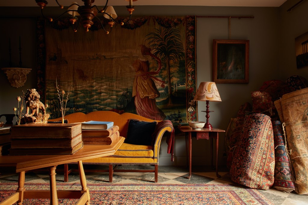 sitting room with yellow sofa and wooden side table, with tapestry depicting a woman in front of the sea in the background and rolled up ornate rugs off to the side