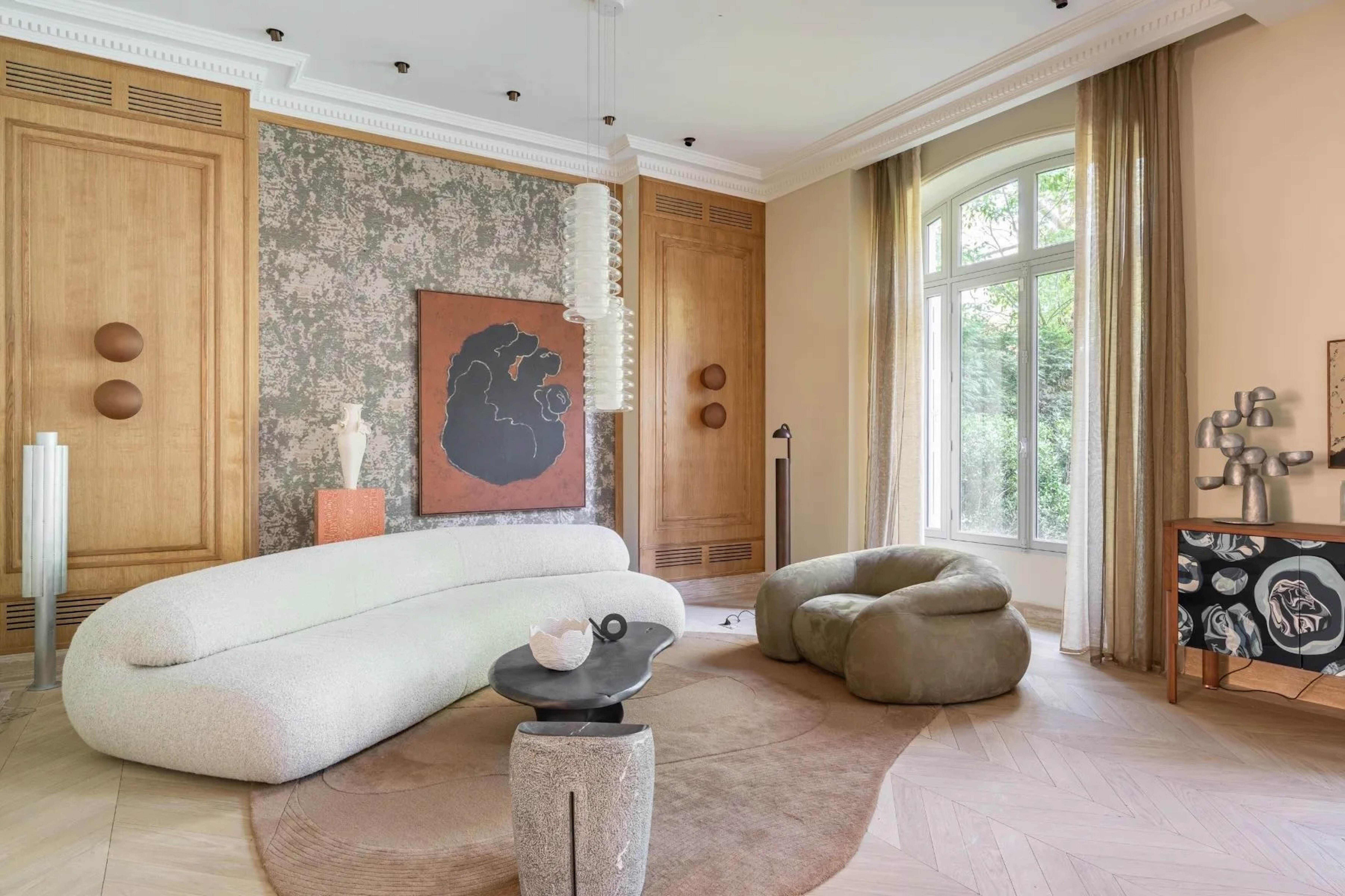 salon in Paris home, featuring modern, curved seating, artistic furniture, including coffee table and console table, with many free standing art pieces and light fixtures in a neutral palette 