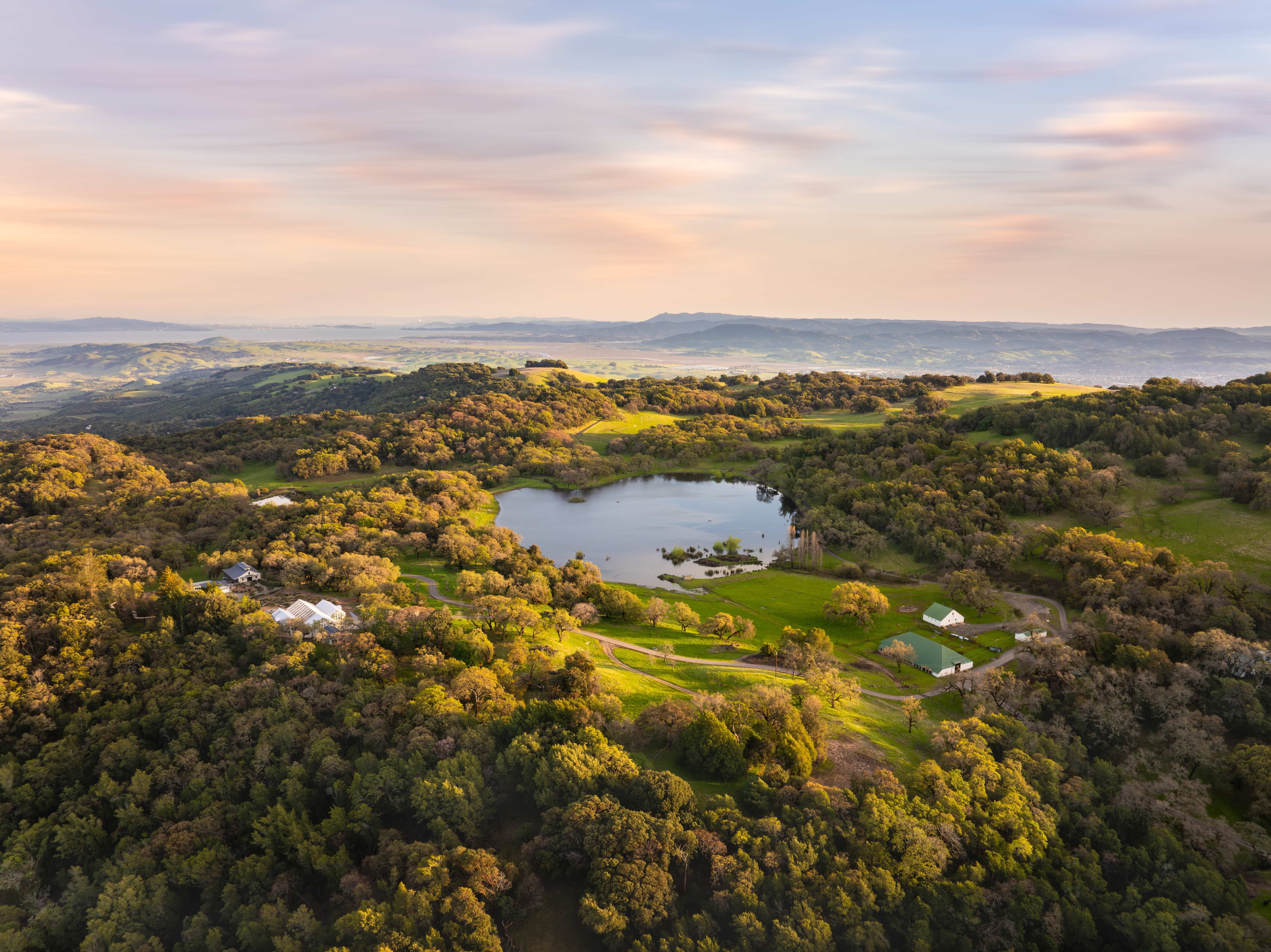 Aerial view of a rural estate featuring a lake, rolling hills, wooded landscapes, agricultural buildings, and distant mountain views under a pastel sky.