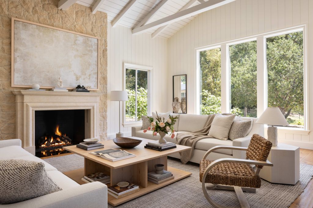 Bright living room with a vaulted white ceiling, stone fireplace, neutral furnishings, large windows, and views of surrounding greenery.