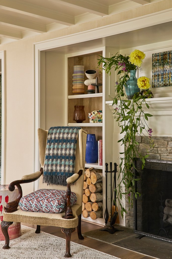 Phillip Thomas designed interior, featuring a chair with cushion and throw near wood-burning fire, with bookshelves in the background featuring various ceramics, books, and wood logs. a trailing plant sits on the mantle