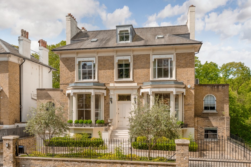 Three-story brick residence with a slate roof, bay windows, and landscaped front garden behind a wrought-iron fence on a sunny day.