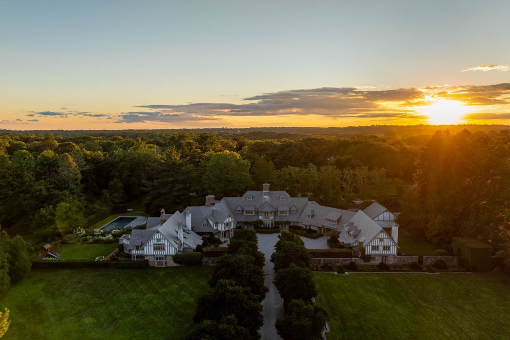aerial shot of grey and white Tudor-style home with pool, tree-lined drive, and pool surrounded by trees, at sunset