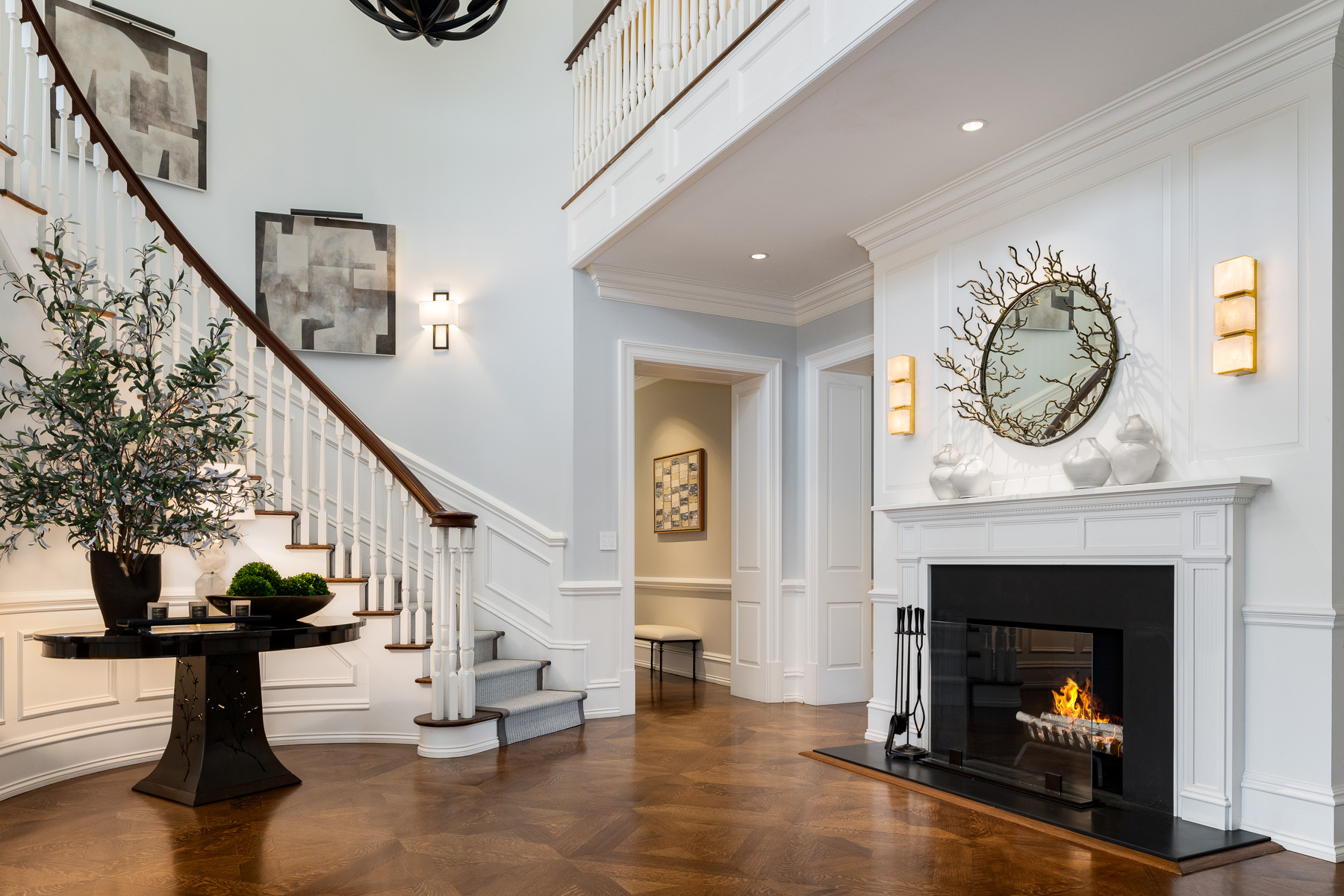 entryway of Greenwich, CT home with parquet floors, featuring a fireplace with white molding, sculptural figures on the mantel, and round mirror with branches stretching outward, and a console table with various plants and a curved staircase in the background