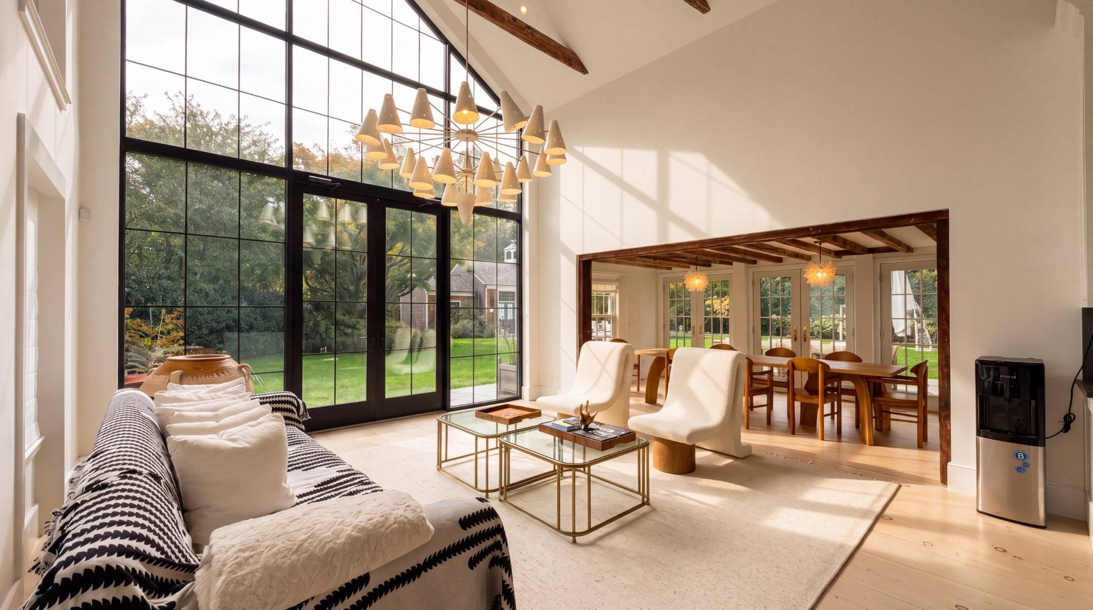 interior of Sag Harbor, NY home, with neutral color palette, including a sofa, two glass and metal coffee tables, and large floor-to-ceiling windows with transparent french doors. Dining room with wood table and chairs and exposed ceiling beams in background