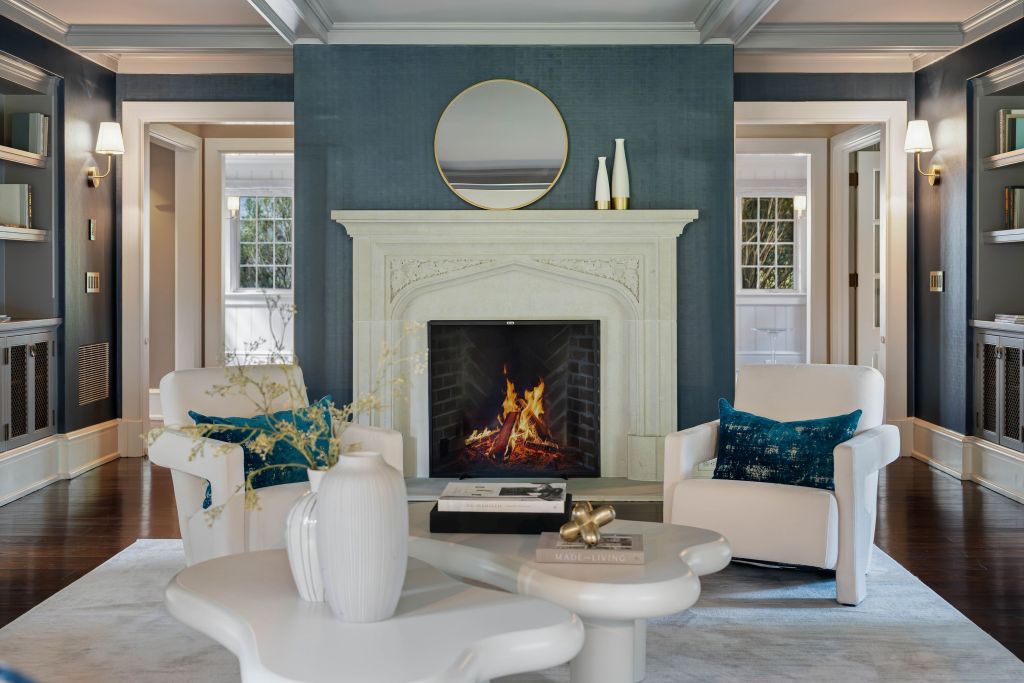 cozy den with blue walls, white abstract coffee table, two white chairs with blue accent pillows, and a roaring marble fireplace in the background