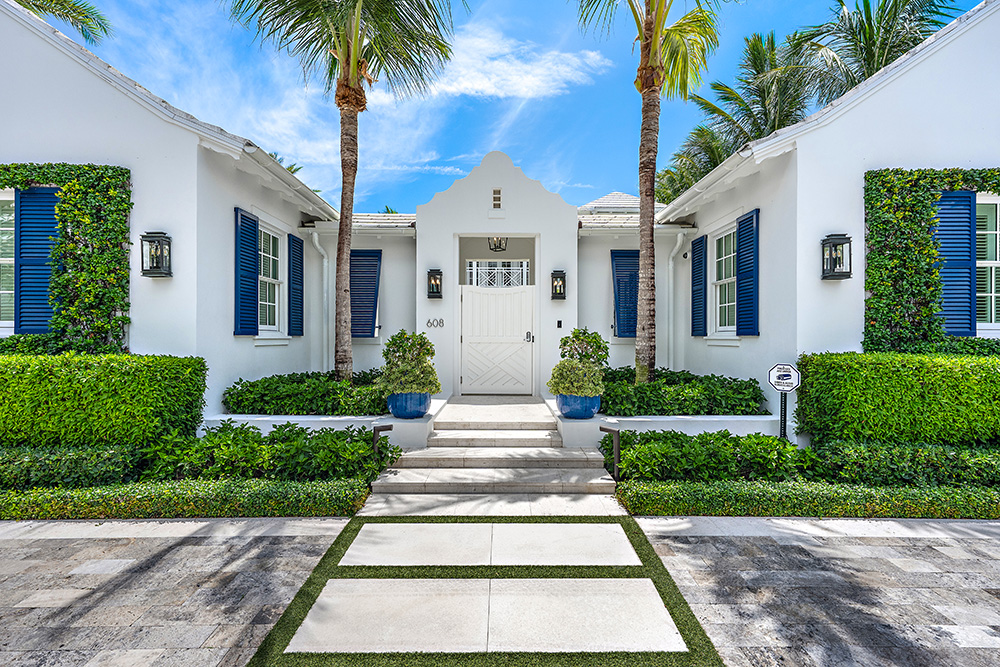 White tropical-style home with blue shutters and palm trees framing a central entryway, surrounded by manicured hedges and a stone walkway under a bright blue sky.