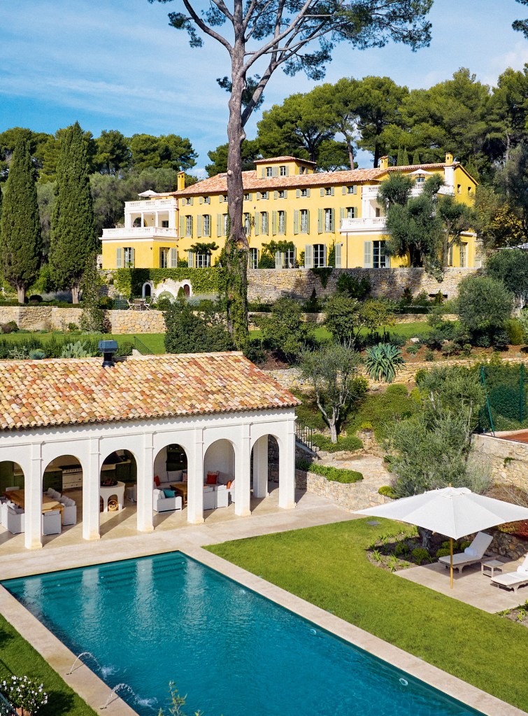 Exterior photo of Chateau Lerins, with pool and covered outdoor seating with terracotta tiles, arched openings, and columns in the foreground. In the background, trees and landscaping frame a yellow chateau with matching terracotta roof tiles, perched atop stone walls
