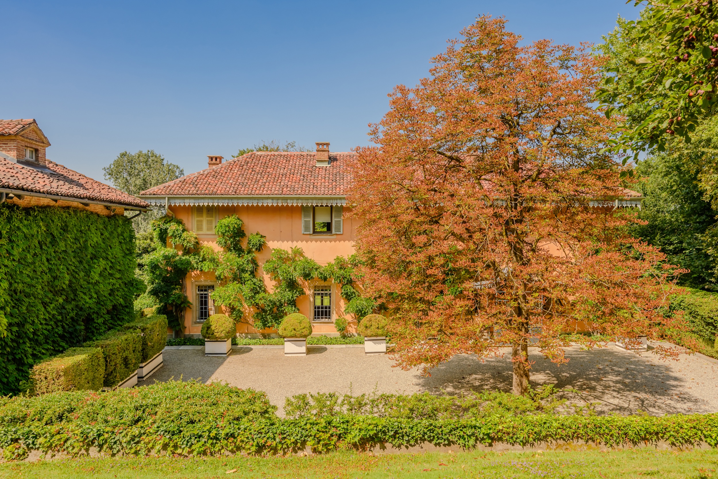 photograph of orange stucco home with terracotta roof tiles in Turin, Italy, featuring lush landscaping, trailing vines, and a mature tree with auburn leaves in the foreground