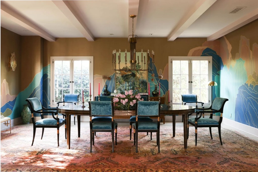 dining room in Los Feliz, CA home with blue velvet chairs, a large floral arrangement on the table, and a blue and brown hand-painted mural on the walls, with a brass chandelier handing from the ceiling and windows letting in natural light in the background