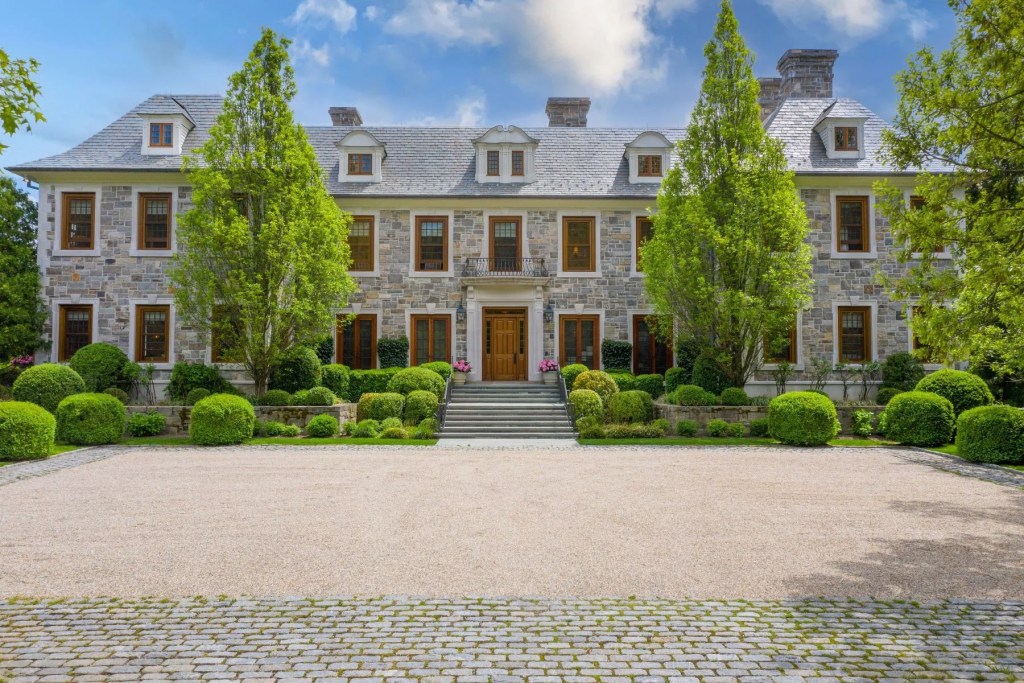 Rear view of a large stone estate home with manicured terraced gardens, broad lawns, and mature trees surrounding the property.