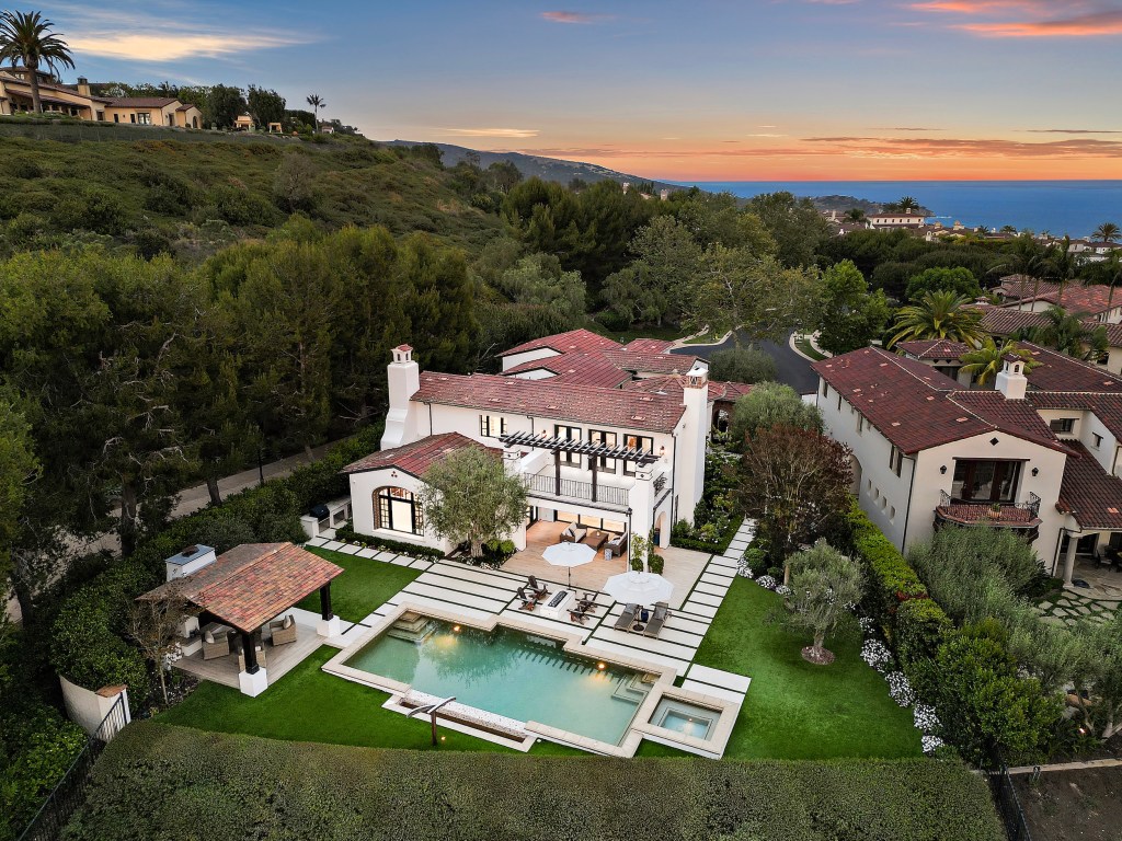 Aerial view of a Mediterranean-style white residence with a red tile roof, swimming pool, spa, and landscaped grounds, surrounded by neighboring homes and coastal hills at sunset.