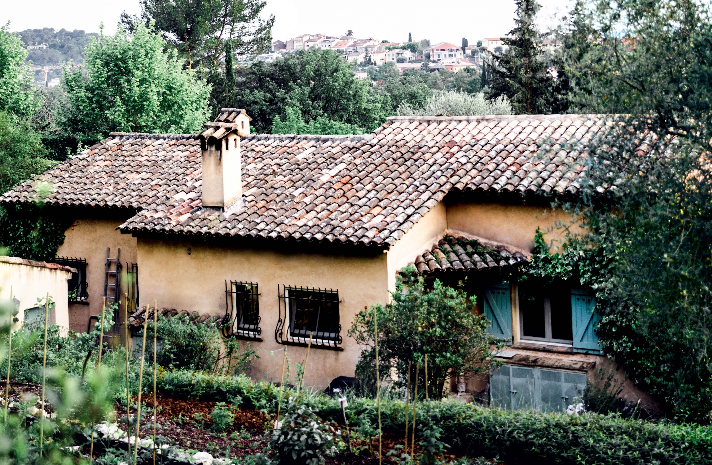 Exterior photo of Julia Child's French country cottage, with tile roof, terracotta stucco walls, blue shutters, and ornate wrought iron window coverings, surrounded by verdant landscaping