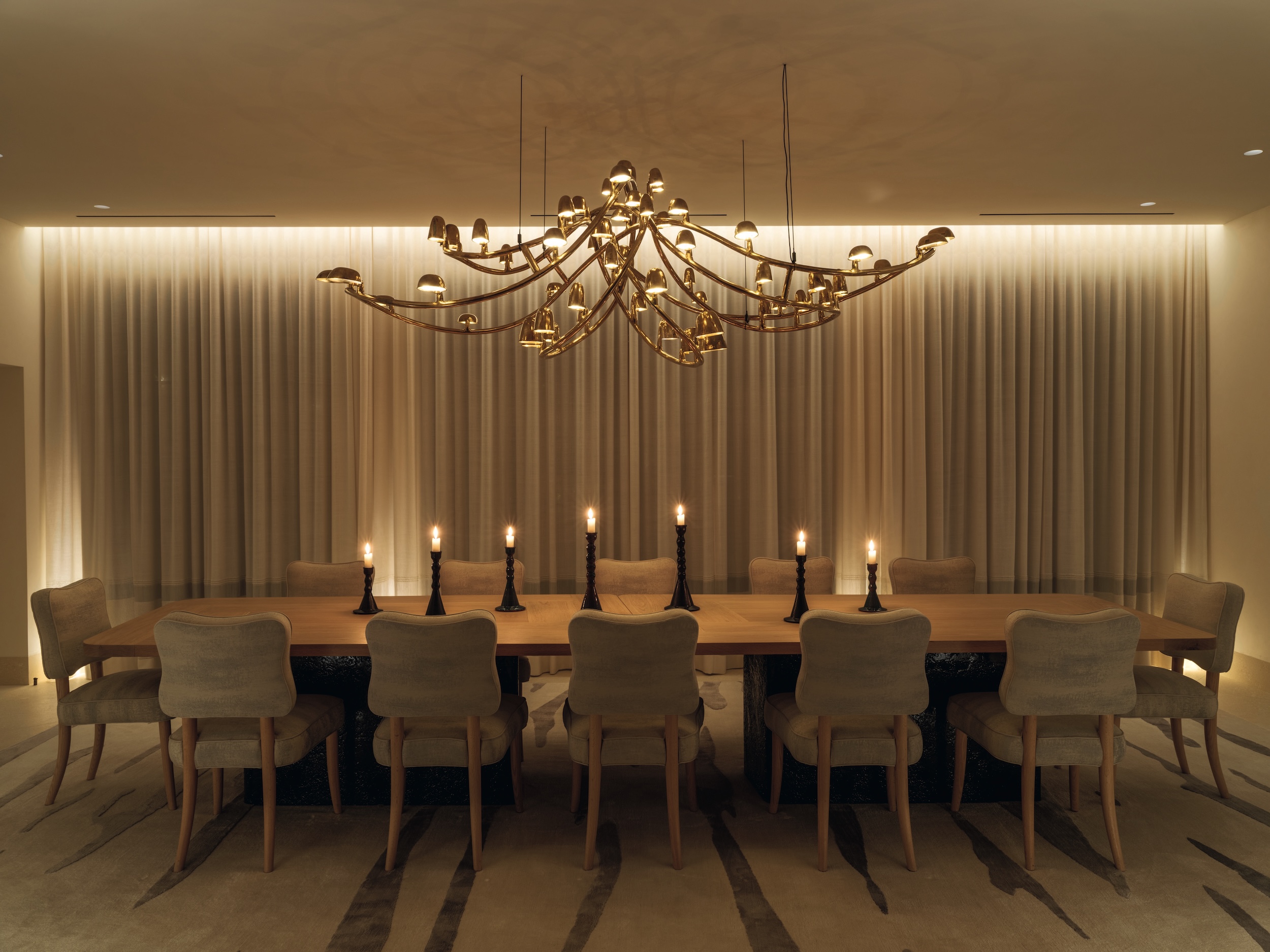 large dining table with 12 plush chairs, 7 black candlesticks with candles burning, and an ornate brass chandelier curving in multiple directions, background with curtained windows