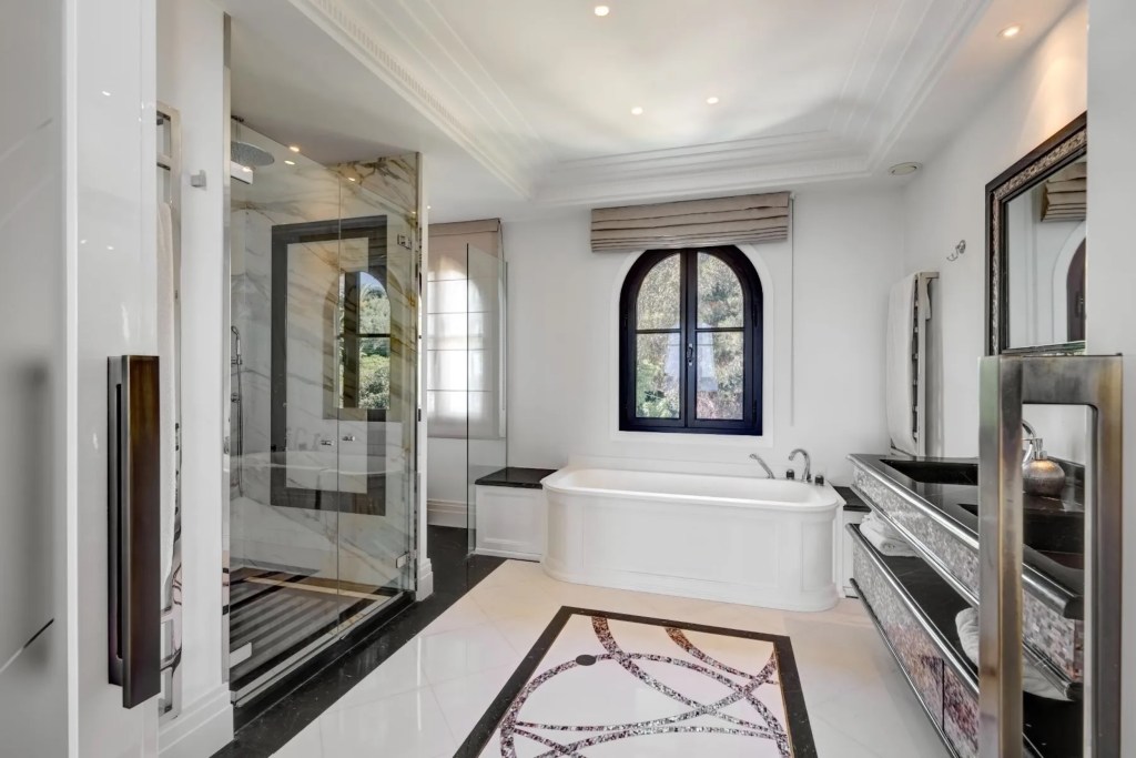 black and white monochromatic bathroom in Provence, with large soaking tub, black and white marble shower with glass door, intricate floor design, and black framed arched window