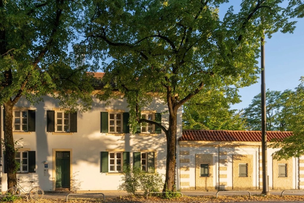 Exterior of white stucco home in Munich, with yellow molding around the windows and doors, green shutters, and tile roof, framed by large trees