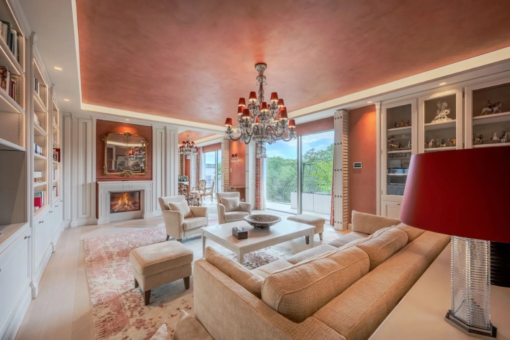 interior of Munich penthouse, with white elegant furnishings and built-in shelving, white fireplace with mirror overhead, and red painted ceilings with doors leading to the balcony beyond