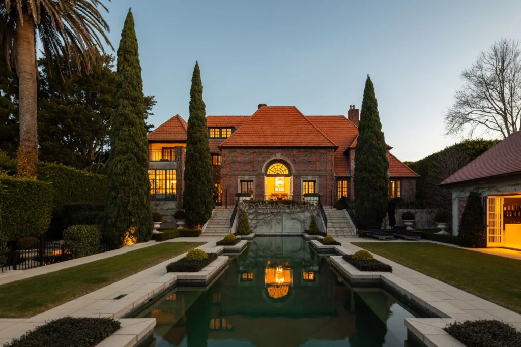 Brick estate residence at dusk with illuminated windows, a formal reflecting pool, symmetrical landscaping, and tall cypress trees framing the entrance.