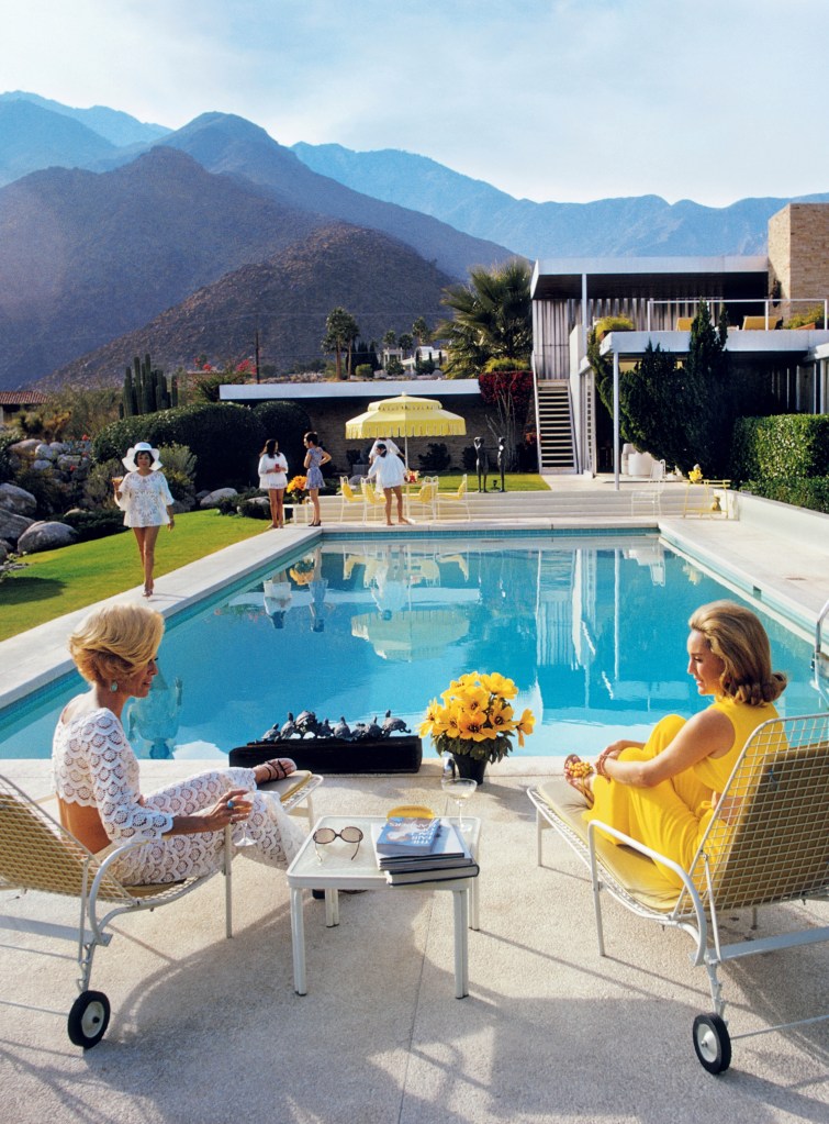 a photo by Slim Aarons from The Kaufmann Desert house in 1970, featuring two women chatting poolside in the foreground. several other ladies, with yellow umbrellas and chairs, and the Palm Springs mid-century home and rolling mountains in the background