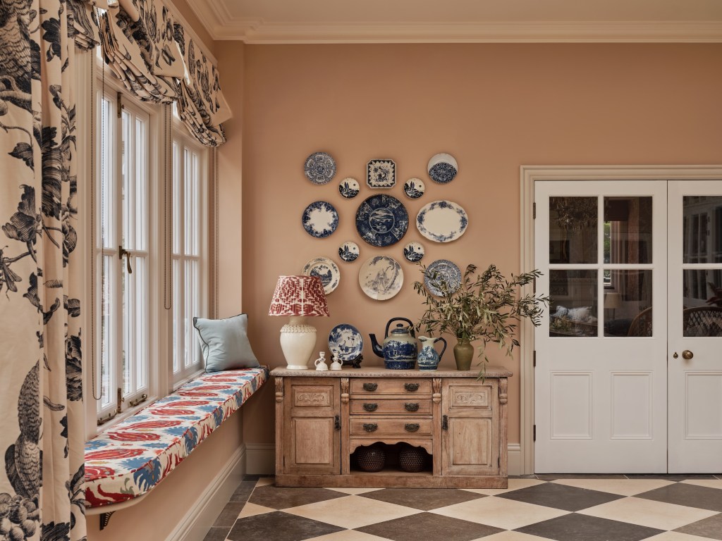 photo of interior designer Rebecca Hughes plate display, situated over an antique console table with drawers and cabinets, featuring other plates and ceramic tea pot, and a window seat with vibrant upholstery and unique patterned window treatments
