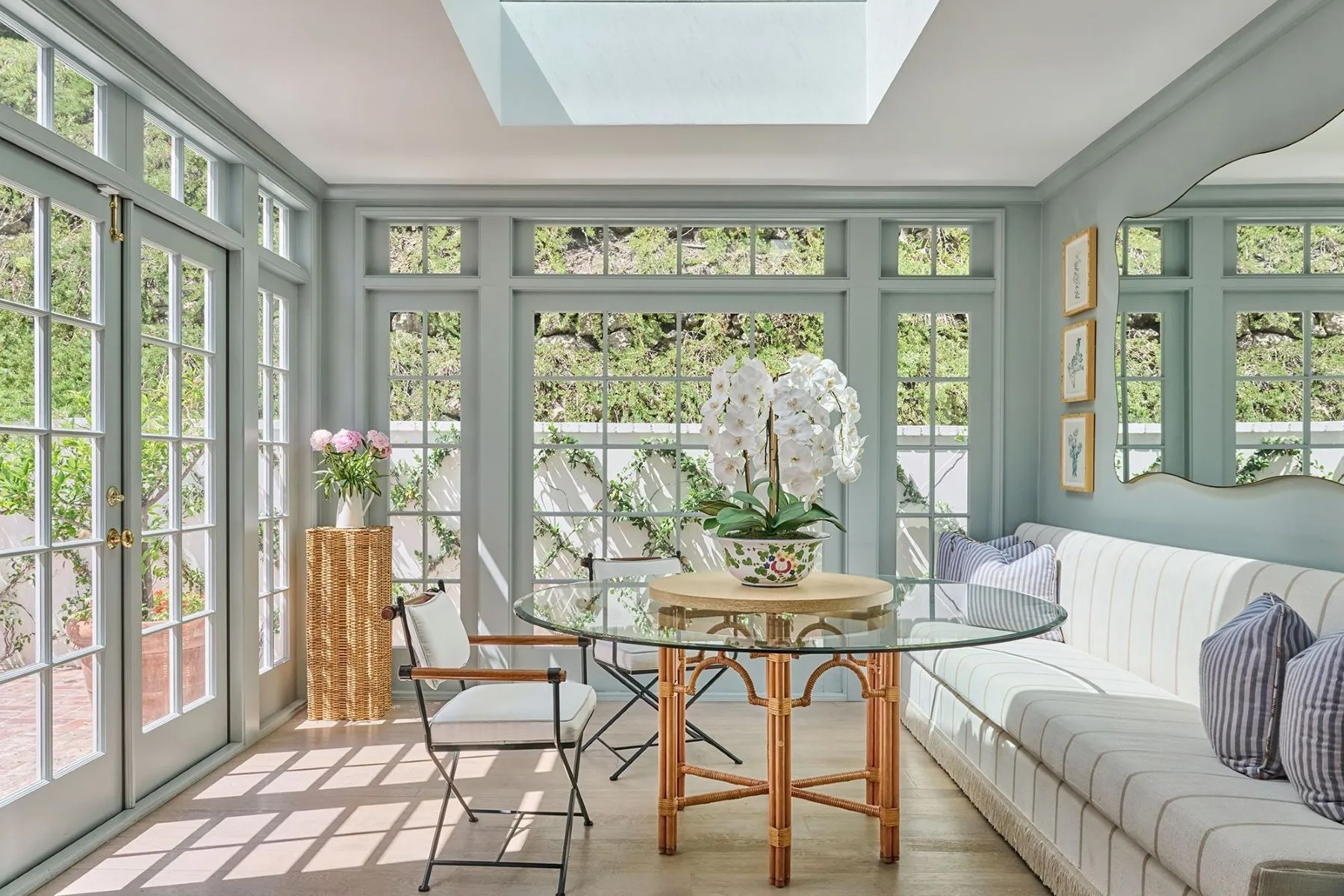 Bright sunroom with pale blue walls, floor-to-ceiling windows, a skylight, a round glass-top table with white orchids, and built-in bench seating overlooking a garden courtyard.