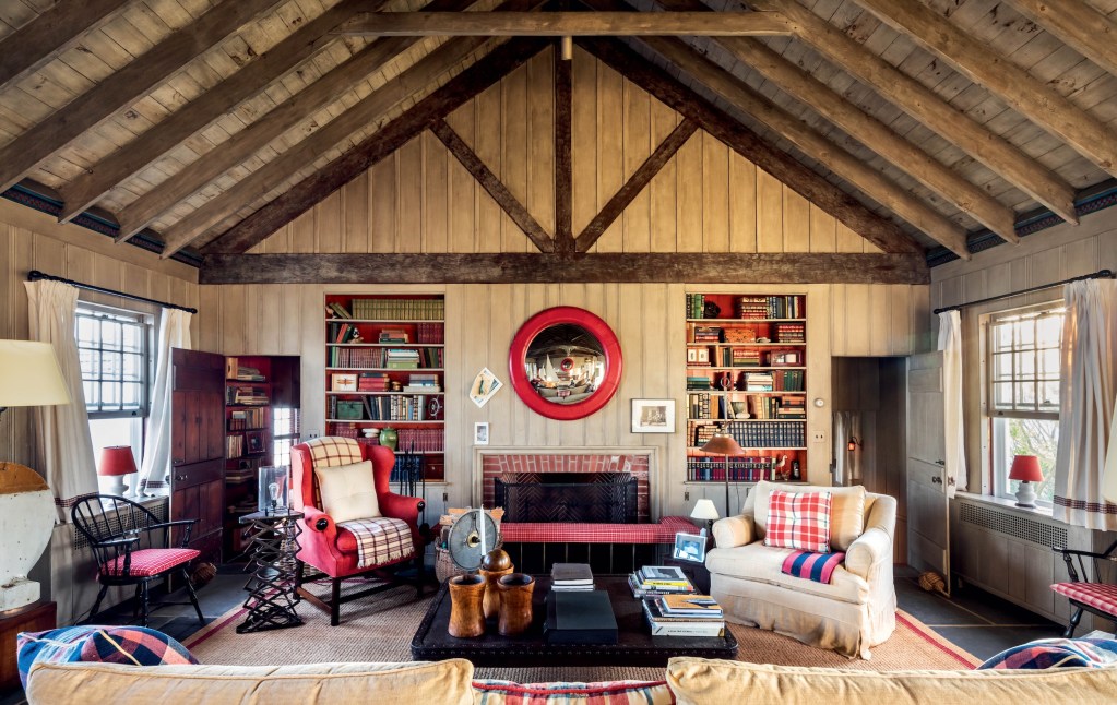 cozy guest house interior of Any Warhol's Montauk home, with plush chairs and sofas situated before a brick fireplace, with exposed wood beam ceiling, bookshelves lined with books, and two open windows and doors