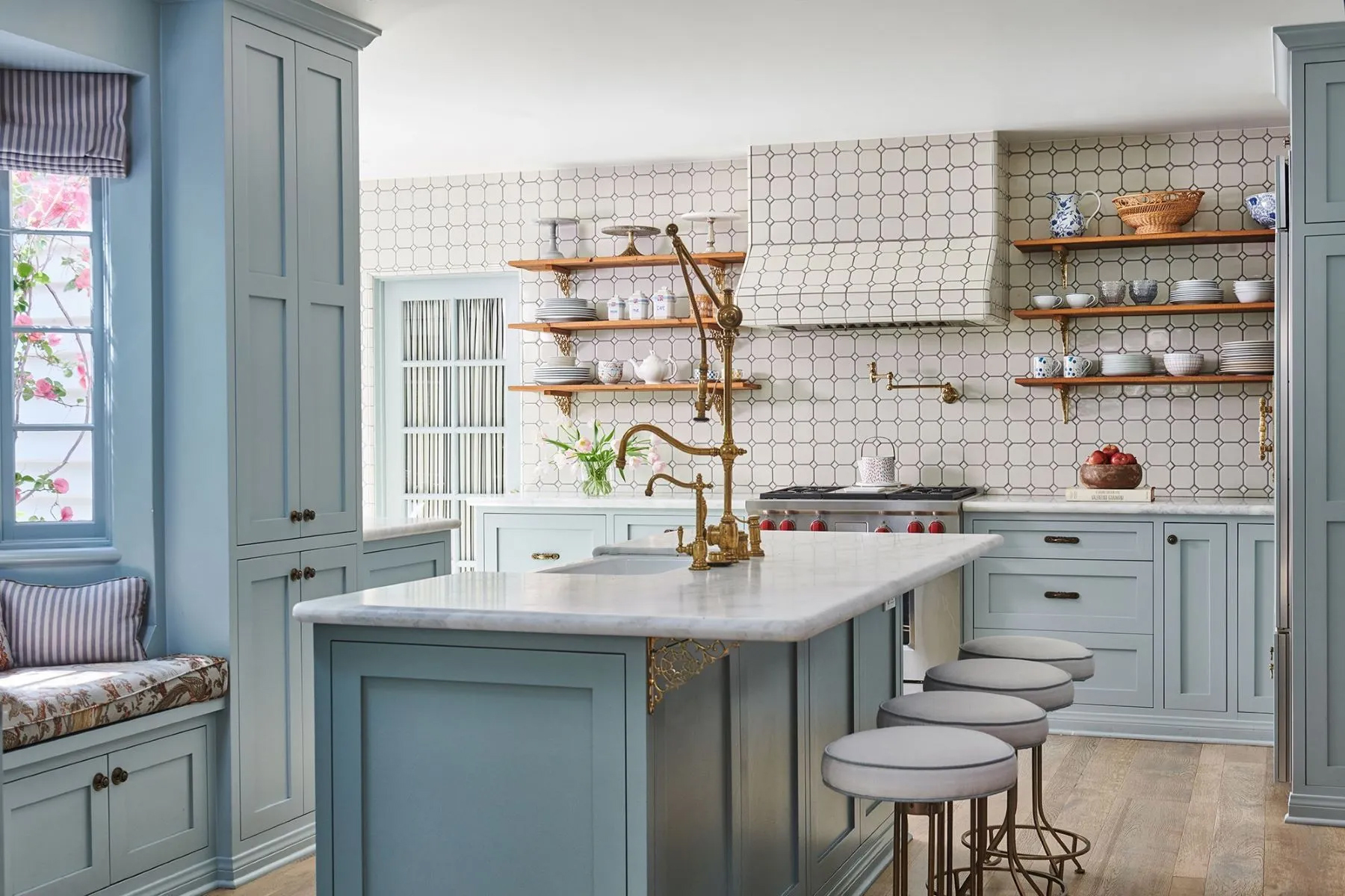 Light blue kitchen with a marble island, brass fixtures, open wood shelving, white geometric tile backsplash, and seating for three at the island.