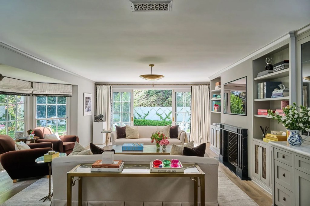Bright, neutral-toned living room with two sofas, armchairs, built-in shelving, a black fireplace, and large French doors opening to a landscaped garden.