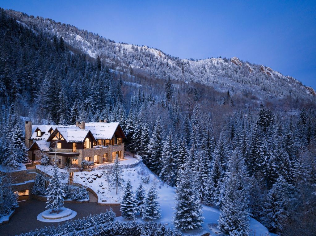 Luxury mountain home nestled among snow-covered evergreen trees, illuminated at dusk against a backdrop of snow-covered mountains and a clear winter sky.